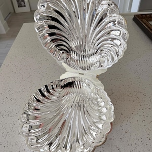 May include: A silver-toned metal clam shell dish with a hinged design that opens to reveal two concave, scalloped interior surfaces. The reflective, polished finish features deep, radiating grooves that mimic the natural texture of a seashell.