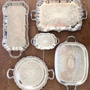 May include: A collection of five silver-toned serving trays with ornate handles and intricate embossed patterns. The set includes one tall rectangular tray, one large rectangular tray, one small oval tray, one round tray, and one large oval tray.