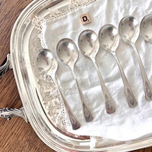 May include: A collection of silver spoons arranged on a silver serving tray with ornate handles. The spoons rest on a white cloth with lace trim. A wooden tile with the letter "D" and the number "2" is also present.