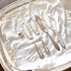 May include: A silver serving tray holds four antique butter knives on a white linen napkin with lace trim. The knives have ornate handles and curved blades. A wooden tile with the letter "F" is also present.