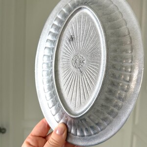 Puede incluir: Plato ovalado de metal plateado con un borde texturizado y un dise&ntilde;o de rayos de sol en el centro. El plato tiene un borde ligeramente elevado y un patr&oacute;n decorativo alrededor del per&iacute;metro. El plato es sostenido por una mano.
