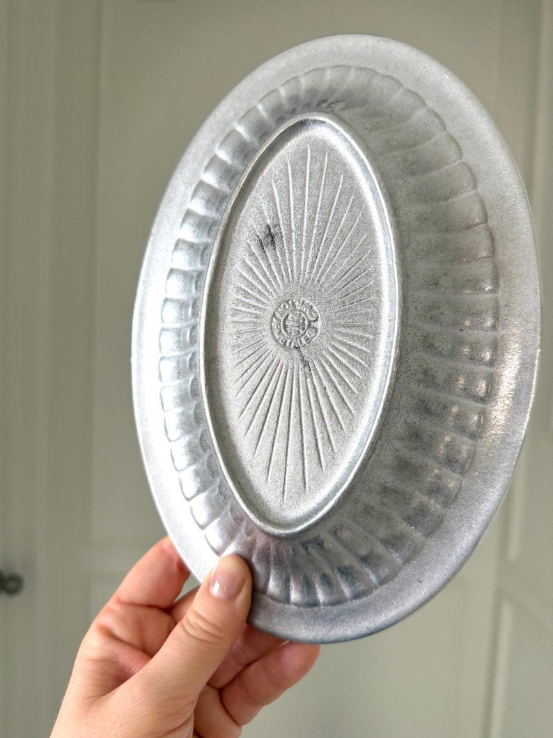 Puede incluir: Plato ovalado de metal plateado con un borde texturizado y un dise&ntilde;o de rayos de sol en el centro. El plato tiene un borde ligeramente elevado y un patr&oacute;n decorativo alrededor del per&iacute;metro. El plato es sostenido por una mano.