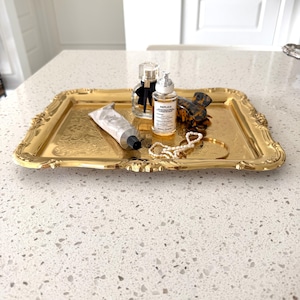 May include: A rectangular gold-toned metal tray with ornate embossed edges. On the tray are a clear glass perfume bottle with a black bow, a smaller clear bottle labeled Replica, a silver tube, a tortoiseshell hair clip, and a pearl necklace.