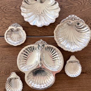May include: A collection of silver-colored serving dishes and trays, including a three-section tray, and several shell-shaped dishes. The pieces are arranged on a wooden surface, showcasing their intricate details and reflective surfaces. The collection includes various sizes.