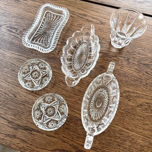 May include: A collection of clear, pressed glass serving dishes and decorative pieces. Includes a rectangular dish, oval trays, a small tumbler, and two round coasters, all with intricate patterns. The pieces are arranged on a wooden surface.