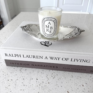 May include: A clear glass candle with a white label reading AMBRE diptyque 34 boulevard saint germain, resting in an ornate silver-toned metal tray atop two stacked hardcover books titled Ralph Lauren A Way of Living and Interior Narratives.