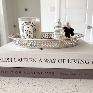 May include: A silver-toned metal tray with an intricate openwork lattice rim, holding a Diptyque Ambre candle and two glass perfume bottles, resting atop two stacked hardcover books titled Ralph Lauren A Way of Living and Interior Narratives.