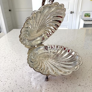 May include: A silver-toned metal serving dish shaped like an open clam shell. The dish features a hinged lid with a decorative handle and is supported by small, ornate feet. The interior and exterior surfaces have a polished, reflective finish.