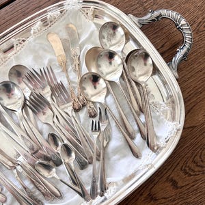 May include: A collection of antique silverware, including spoons, forks, and butter knives, arranged on a silver serving tray. The silverware has ornate detailing and is displayed on a white, lacy cloth. The tray has a decorative handle.