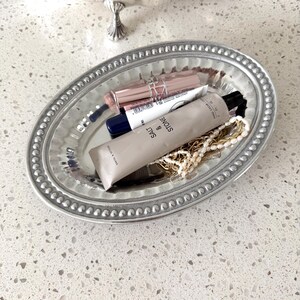 Puede incluir: Una bandeja ovalada plateada con borde de cuentas contiene productos de belleza y joyas. Los art&iacute;culos incluyen barras de labios, un tubo de crema de manos y un collar. Otra bandeja plateada es visible en el fondo.