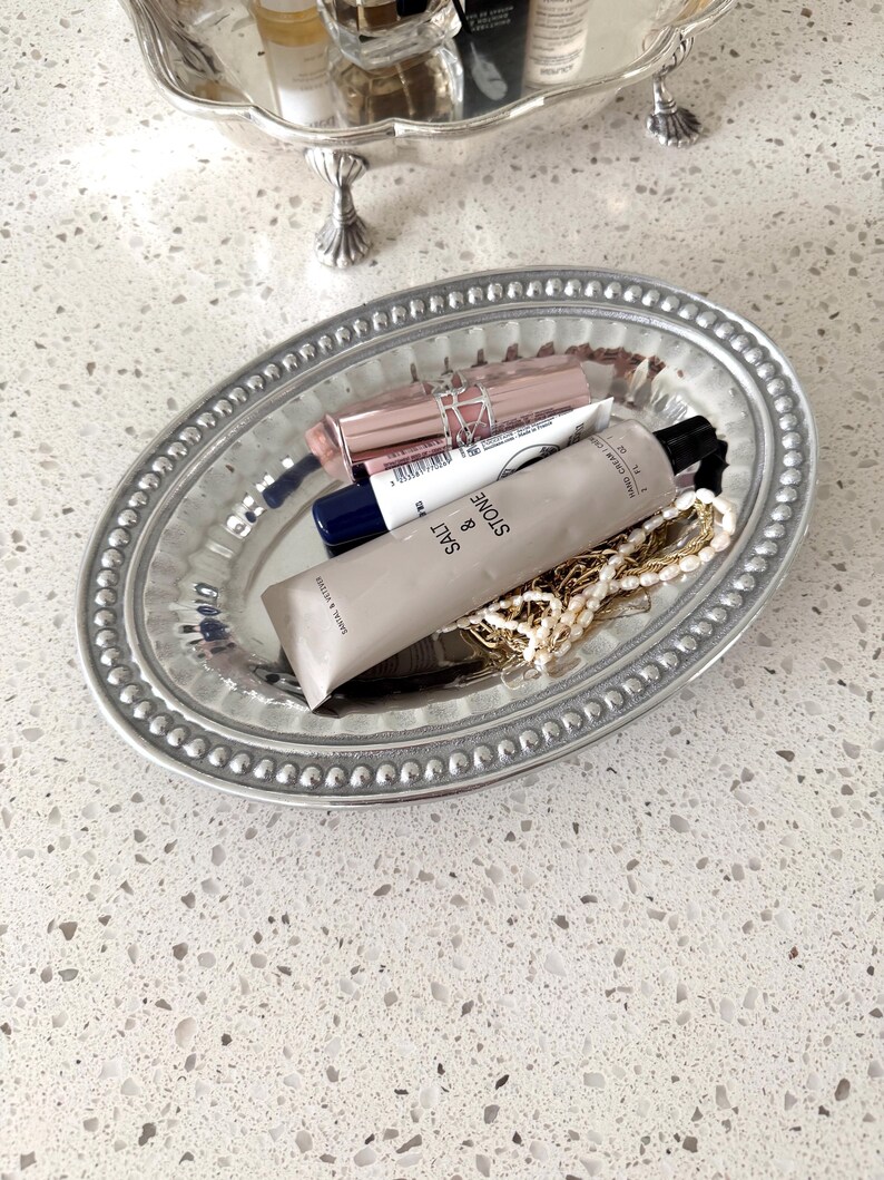 Puede incluir: Una bandeja ovalada plateada con borde de cuentas contiene productos de belleza y joyas. Los art&iacute;culos incluyen barras de labios, un tubo de crema de manos y un collar. Otra bandeja plateada es visible en el fondo.