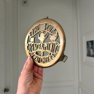 May include: A round brass trivet featuring an openwork design with a teapot, a sugar bowl, and a creamer. The text reads THE CUP THAT CHEERS and PEACE & PLENTY. The metal has a warm, aged patina with visible dark oxidation in the recessed areas.