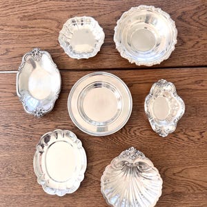 May include: A collection of seven silver-toned metal dishes and bowls of various shapes and sizes. The set includes round, oval, and shell-shaped pieces, some featuring decorative embossed edges and smooth, reflective surfaces for serving.