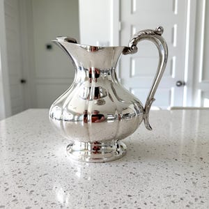May include: A silver-colored pitcher with a bulbous body and a decorative handle. The pitcher has a fluted design and sits on a small, round base. The handle features an ornate, leaf-like detail. The pitcher is reflective, showing the surrounding environment.