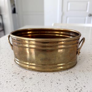 May include: An oval-shaped brass basin featuring a polished metallic finish, a smooth rim, and two small, curved handles attached to the sides. The vessel has a deep interior and a rounded base, showcasing a warm, golden-toned surface.