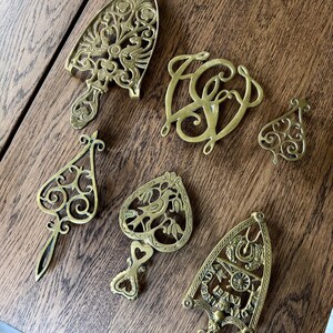 May include: A collection of antique brass trivets and decorative pieces. The items include ornate designs, such as a monogram, a spade shape, and iron-shaped trivets with intricate patterns. The brass pieces are displayed on a wooden surface.