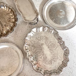 May include: A collection of four silver-toned serving trays and platters. The set includes a shell-shaped dish, an elongated tray with handles, a round platter with an etched center, and a large oval tray featuring an intricate embossed border.