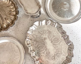 May include: A collection of five silver-toned metal serving trays and platters. The set includes a shell-shaped dish, a long rectangular tray with handles, two round plates with ornate engraved borders, and one large oval platter with a scalloped edge.