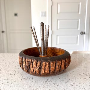 May include: A wooden nut bowl with a bark-like exterior and a dark brown interior. It contains a silver nutcracker and several nut picks. The bowl sits on a speckled white countertop.