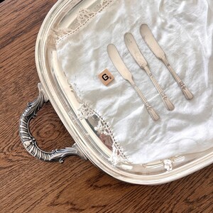 May include: A silver serving tray with ornate handles holds a white cloth with three butter knives. The tray has a decorative edge and is set on a wooden surface. A small wooden tile with the letter "G" is also on the cloth.