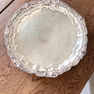 May include: A silver-colored, round serving tray with an ornate, embossed floral design. The tray has a scalloped edge with decorative floral accents. The tray is displayed on a wooden surface.