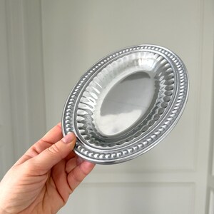 Puede incluir: Un peque&ntilde;o plato ovalado de color plateado con un borde de cuentas y un interior texturizado. El plato se sostiene en una mano, mostrando su forma y dise&ntilde;o. El plato es probablemente para servir porciones peque&ntilde;as o para uso decorativo.