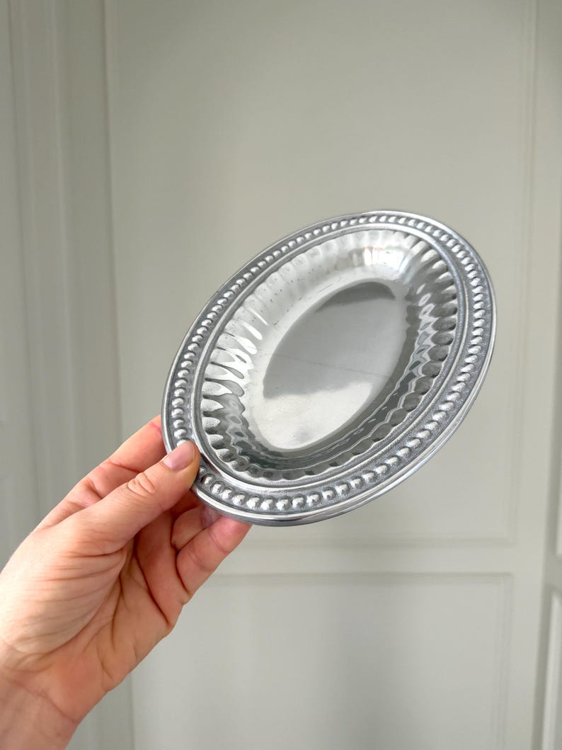 Puede incluir: Un peque&ntilde;o plato ovalado de color plateado con un borde de cuentas y un interior texturizado. El plato se sostiene en una mano, mostrando su forma y dise&ntilde;o. El plato es probablemente para servir porciones peque&ntilde;as o para uso decorativo.