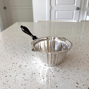 May include: A silver-colored sauce boat with a dark brown handle. The boat has a scalloped edge and a pouring spout. The handle is attached to the side of the boat. The sauce boat is sitting on a white countertop.