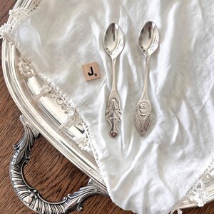 May include: Two ornate silver spoons rest on a white, lace-trimmed cloth, placed on a silver serving tray. A wooden tile with the letter "J" is next to the spoons. The tray sits on a dark wooden surface.