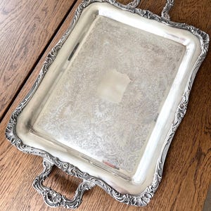 May include: A rectangular silver-toned serving tray featuring ornate, raised scrollwork along the edges and two decorative handles. The central surface displays an intricate, engraved floral pattern with a smooth, blank rectangular area in the center.
