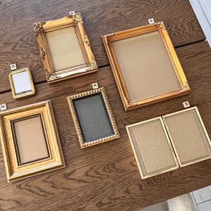 May include: A collection of gold-toned picture frames in various sizes and styles, including ornate and simple designs. The frames are empty, with visible backing, and arranged on a wooden surface. Some frames are rectangular, and one is a small, square shape.
