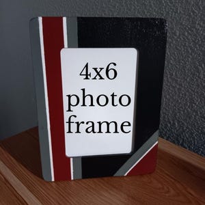 May include: A rectangular photo frame with a 4x6 inch opening. The frame is painted with a design of black, gray, and burgundy stripes. The frame is sitting on a wooden surface.