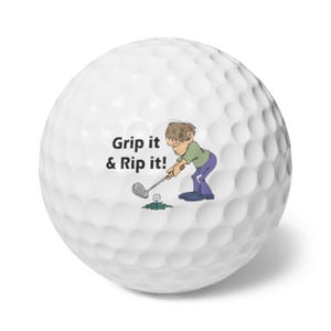 Könnte beinhalten: Weißer Golfball mit einer genoppten Oberfläche. Der Ball zeigt eine Cartoon-Illustration eines Golfspielers, der einen Schläger schwingt, mit dem Text "Grip it & Rip it!" in Schwarz. Der Golfer trägt ein grünes Hemd und eine lila Hose.