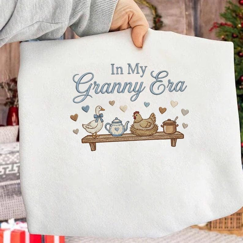 May include: White fabric featuring the embroidered text "In My Granny Era" in blue script. Below, a duck with a blue bow, a teapot, a hen, and a pot are on a wooden shelf, with heart accents.