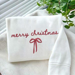 Könnte beinhalten: Ein weißes Sweatshirt mit dem Schriftzug "merry christmas" in roter Schrift und einer roten Schleifen-Grafik. Das Sweatshirt ist gefaltet, ein Ärmel ist sichtbar. Der Hintergrund umfasst eine weiße Oberfläche und grüne Blätter.