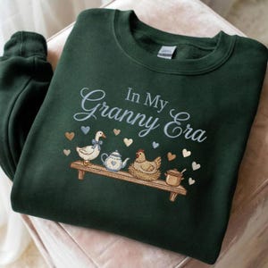May include: Dark green sweatshirt featuring the embroidered text "In My Granny Era." The design includes a goose, teapot, chicken, and hearts. The sweatshirt is folded on a light pink surface.