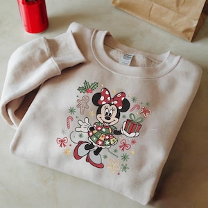 Peut inclure: Un sweat-shirt beige avec une broderie de Minnie Mouse sur le thème de Noël. Minnie Mouse porte un nœud rouge et une robe festive, tenant un cadeau. Le motif comprend des cannes en sucre, des bonhommes en pain d'épices et du houx.
