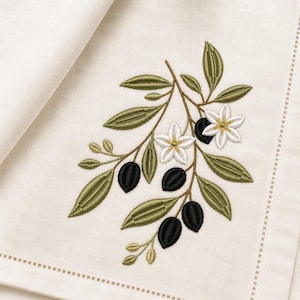 Può includere: Tovagliolo di lino color crema con un ricamo a ramo d'ulivo. Il ramo presenta foglie verdi, olive nere e fiori bianchi con centri dorati. Il tovagliolo ha un bordo cucito.