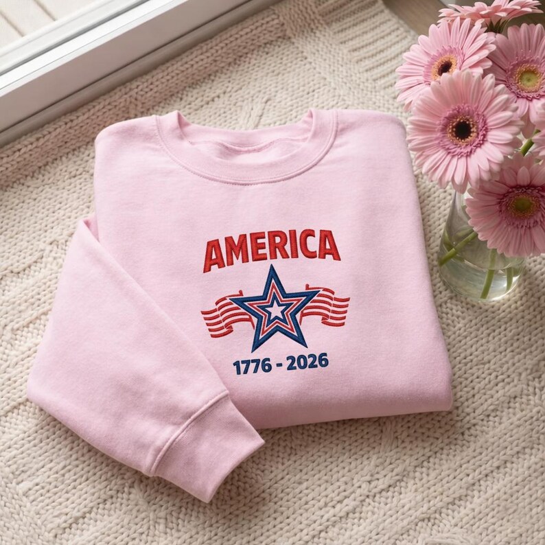 May include: A light pink sweatshirt with the word "AMERICA" in red, a blue star with a white star in the center, and red and white stripes. The text "1776-2026" is below the star. The sweatshirt is folded on a white knitted surface.
