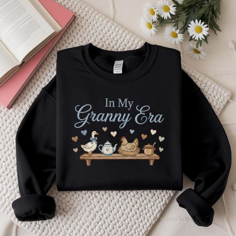 May include: Black sweatshirt featuring the text "In My Granny Era" in light blue script. The design includes a shelf with a duck, teapot, chicken, and pot, along with blue and brown hearts.