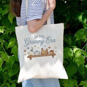 May include: Cream tote bag featuring the embroidered text "In My Granny Era." The design includes a duck, teapot, chicken, and pot on a wooden shelf, with heart accents. The bag has cream-colored handles.