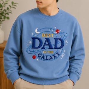 Könnte beinhalten: Hellblaues Sweatshirt mit der gestickten Aufschrift "BEST DAD in the GALAXY". Das Design umfasst einen Halbmond, Planeten und Sterne. Das Sweatshirt ist ein Rundhals-Stil.