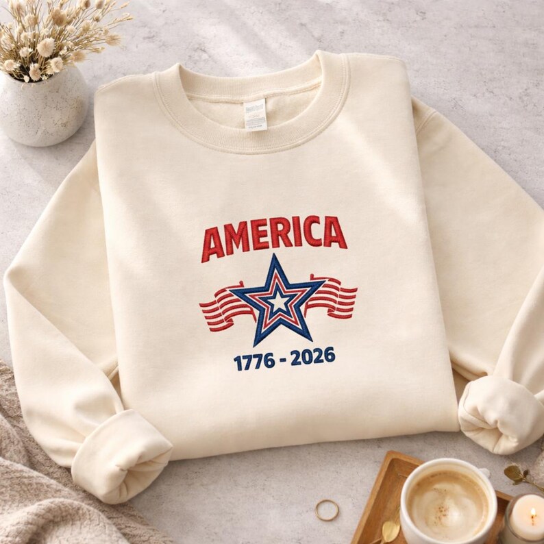 May include: Cream-colored sweatshirt with the word "AMERICA" in red, a blue and white star, and the years "1776-2026" in blue. The design includes red and white striped flags. The sweatshirt is on a light-colored surface.