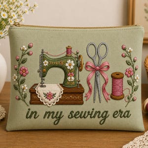 Puede incluir: Un estuche verde claro con cremallera y bordado de máquina de coser, tijeras y carrete de hilo. La frase "in my sewing era" está bordada debajo. Acentos florales y un lazo rosa completan el diseño.