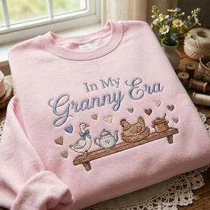 May include: A pale pink sweatshirt featuring the embroidered words "In My Granny Era." The design includes a shelf with a duck, teapot, chicken, and pot. The design includes heart shapes in various colours.