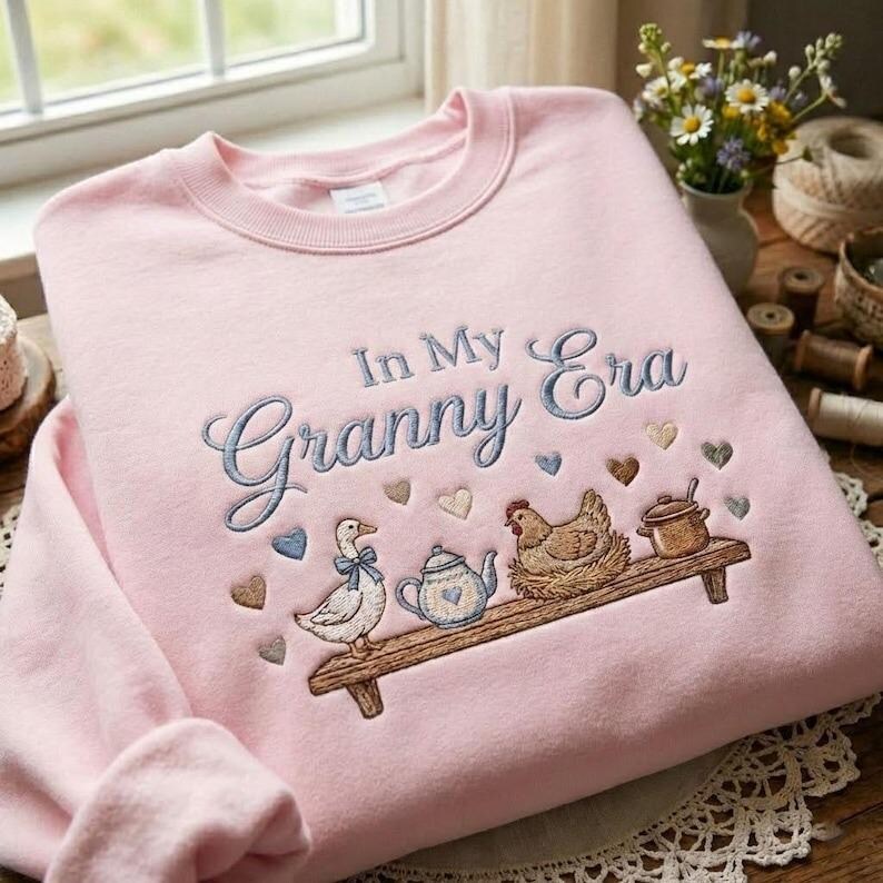 May include: A pale pink sweatshirt featuring the embroidered words "In My Granny Era." The design includes a shelf with a duck, teapot, chicken, and pot. The design includes heart shapes in various colours.