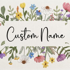 May include: An embroidered design featuring a rectangular space with the words "Custom Name" in black script. The rectangle is framed by colorful embroidered flowers and greenery, including pink, yellow, blue, and white blooms, set against a cream-colored background.