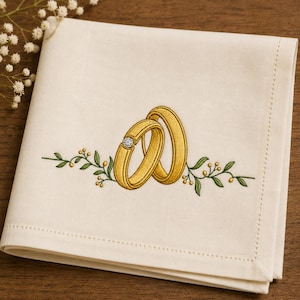 May include: Ivory-colored cloth napkin with embroidered gold wedding rings, one with a diamond. Green and yellow floral embroidery accents the rings. The napkin is folded and has a stitched border.