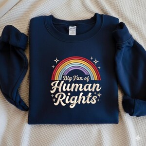 Könnte beinhalten: Marineblaues Sweatshirt mit einem Regenbogen-Design und dem Text "Big Fan of Human Rights". Der Regenbogen ist mehrfarbig, und der Text ist in einer weißen, stilisierten Schriftart gehalten. Das Sweatshirt ist aus weichem Stoff.