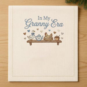 May include: Cream fabric with the embroidered phrase "In My Granny Era". Below, a shelf displays a goose, teapot, chicken, and pot. The design is completed with hearts and a dotted border.